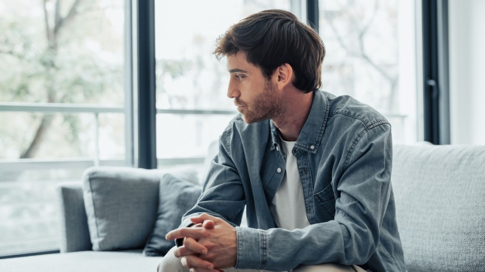 Thoughtful serious man sitting on sofa alone at home, lost in thoughts, thinking about problem solving, feeling lonely, making important decision, having psychological