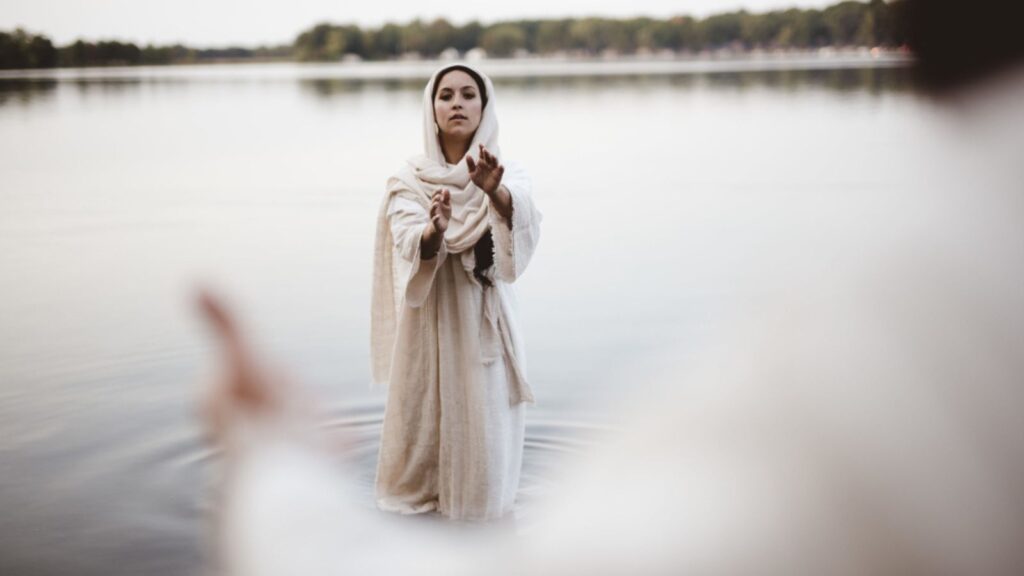 A female wearing a biblical gown while standing in the water and reaching towards Jesus Christ