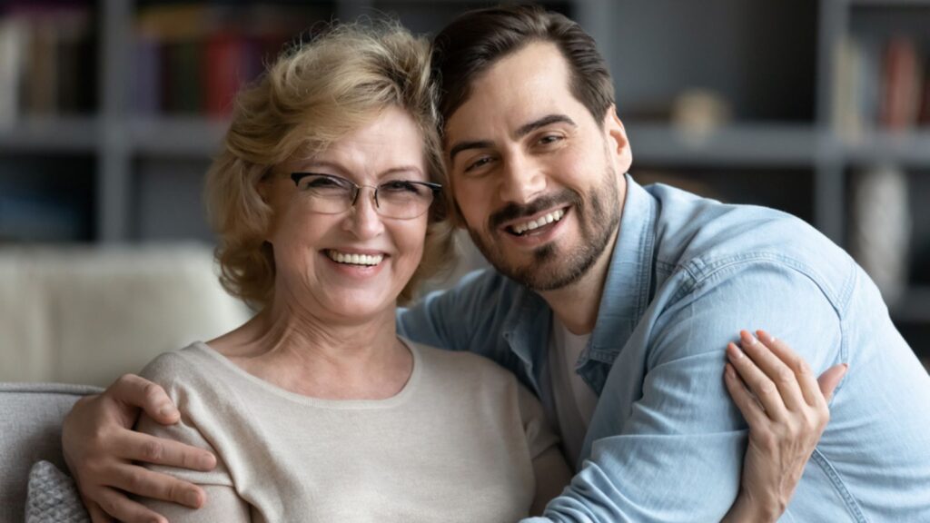 smiling man embracing affectionate older mother in glasses, enjoying sweet tender time indoor, spending weekend