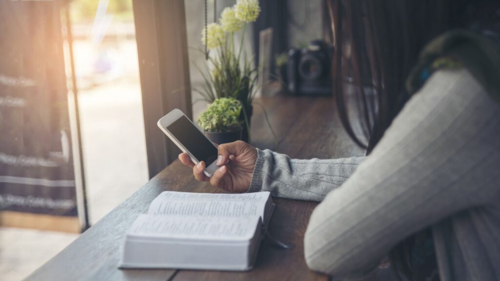 Bible Study concept. Sad Christian woman read bible at coffee shop, send message by mobile phone. Person side view, hold smartphone. Asian student
