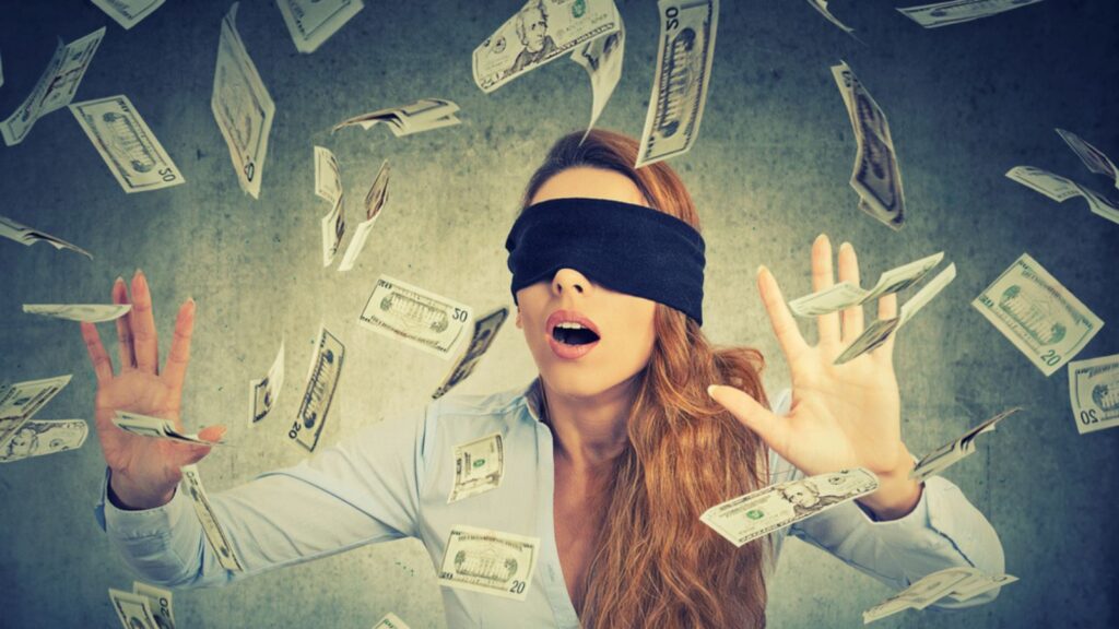 Blindfolded young entrepreneur businesswoman trying to catch dollar bills banknotes flying in the air on gray wall background. Financial corporate success or