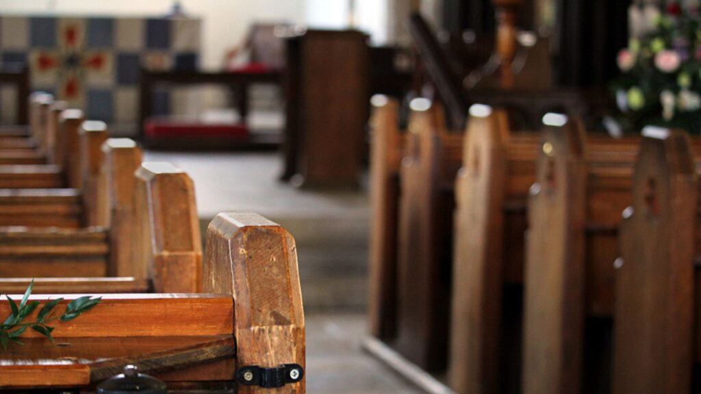 Close up of the end of a row of wooden pews in a traditional stone built church, with the altar in out of focus background