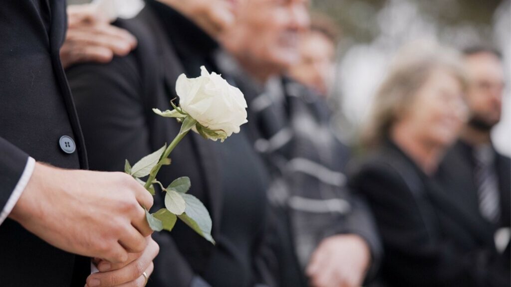Hands, rose and a person at a funeral in a cemetery in grief while mourning loss at a memorial service. Death, flower and an adult in a suit at a graveyard in a crowd