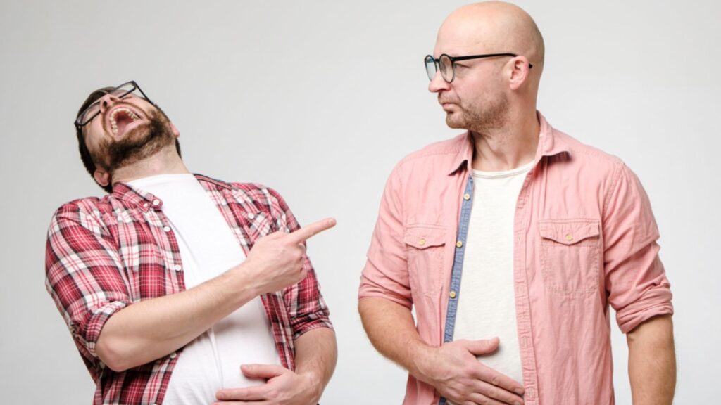 One laughs out loud, holds on to stomach and points a finger at another person who is upset by the evil joke of his friend