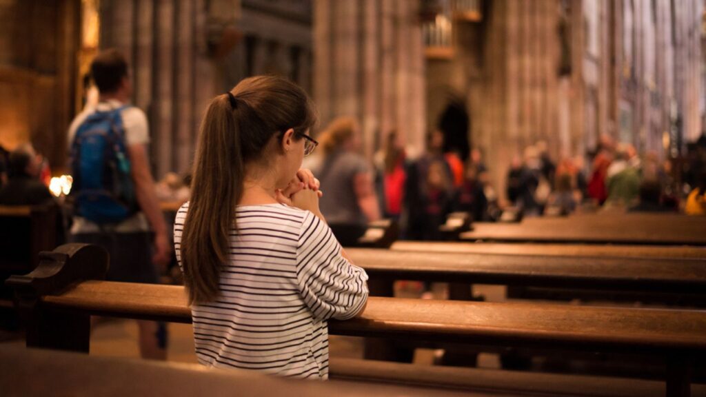 Girl in a church standing on her knees. Young religious woman in glasses with long dark hair, praying in catholic cathedral.,church