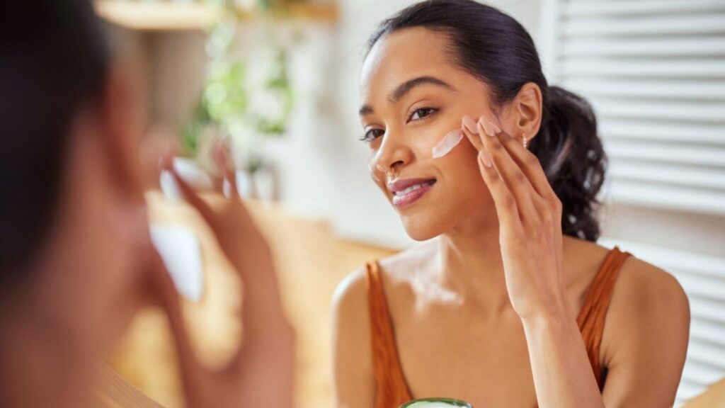 Woman caring of her beautiful skin on the face standing near mirror in the bathroom. Mexican woman applying moisturizer on her face at home. Multiethnic girl holding little green j